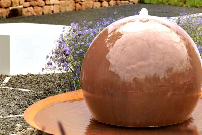Cortenstahl-Kugelbrunnen mit Schale in einem Blumenbeet