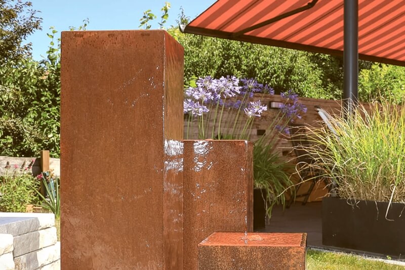 dreisaeuliger-cortenstahlbrunnen-auf-einer-terrasse