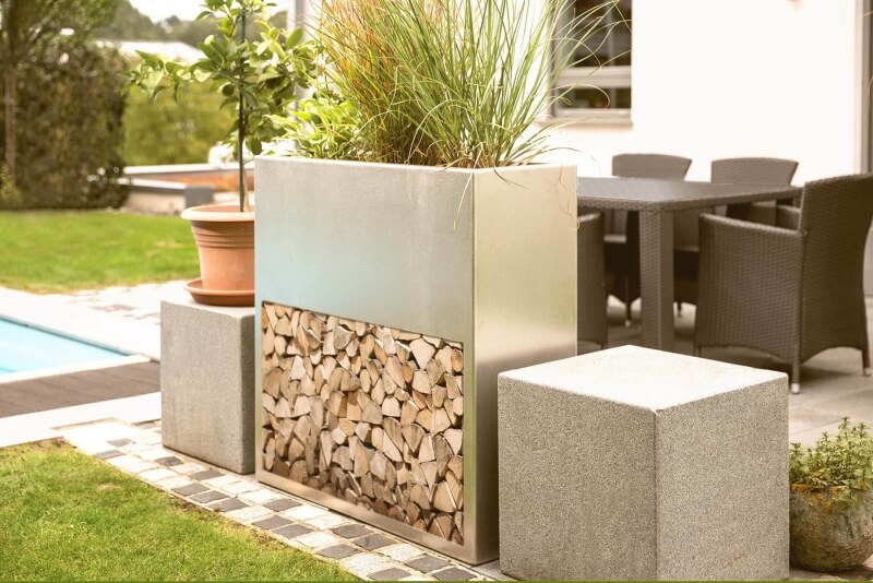 Kaminholzlager aus Edelstahl befüllt mit Holz mit integriertem Pflanzkasten zwischen zwei quadratischen Elementen im Garten