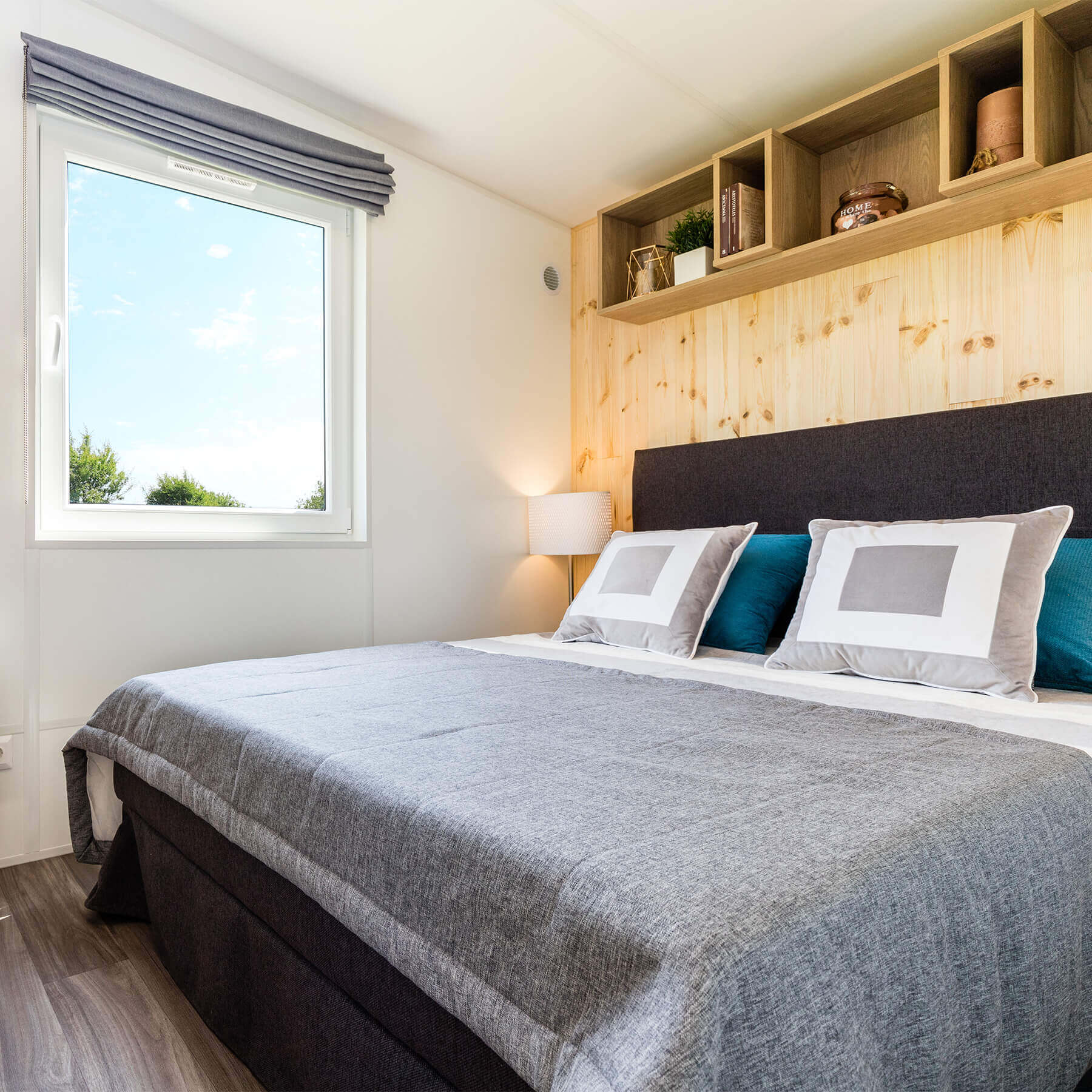 Komfortabel eingerichteter Schlafbereich mit Bett, Nachttisch, Hängeregal und Raffrollo am Fenster